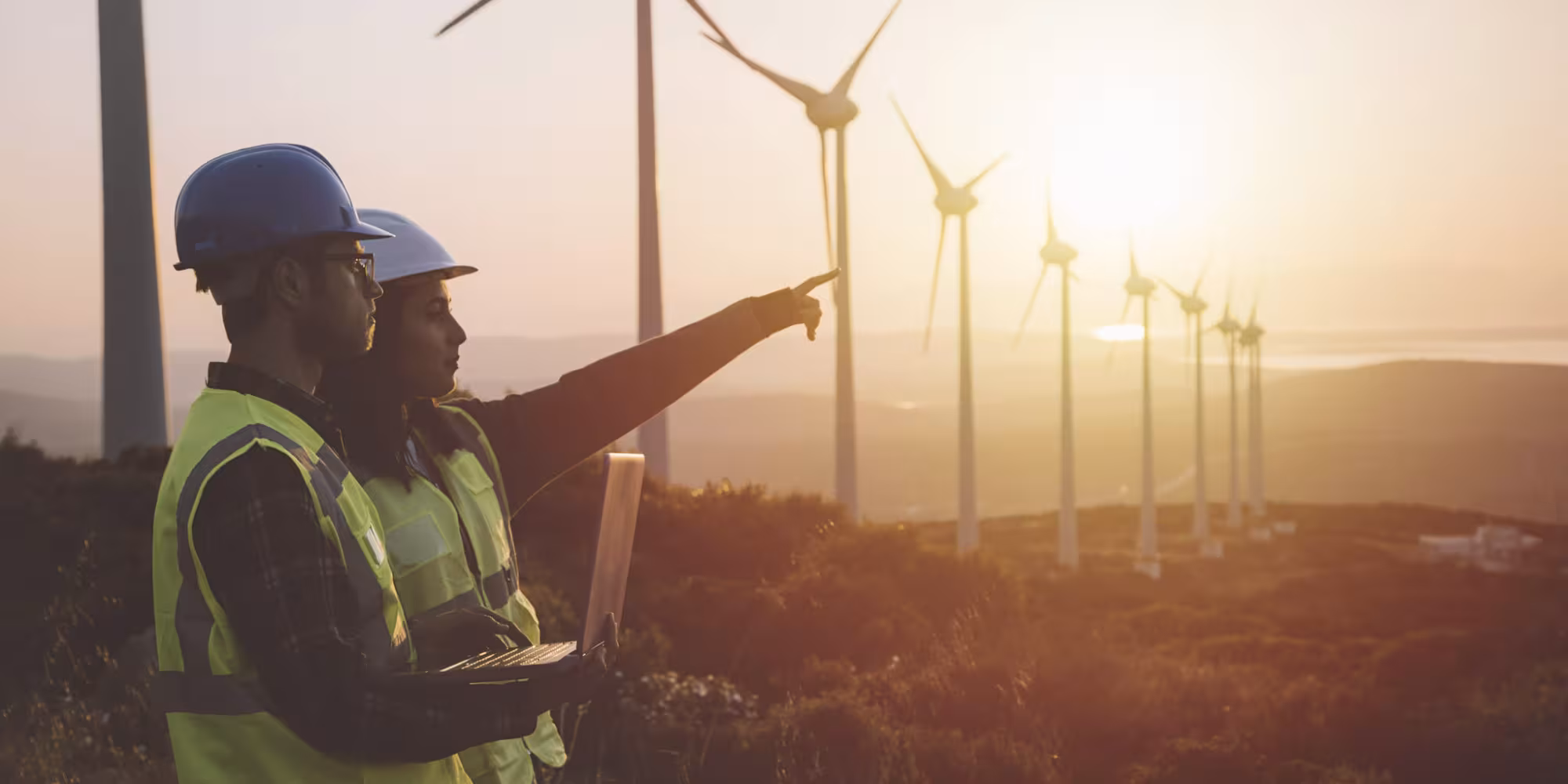 Two people in hi-vis standing in front of a row of wind turbines with the sun setting behind them