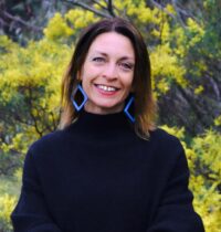 A smiling photo of Sara Bice posing in front of a wattle bush. She is wearing a black turtleneck and big blue diamond shaped earing.