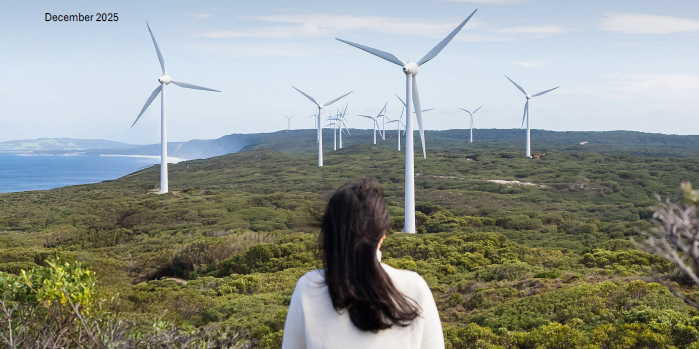 Woman and child on a green mountain side with their backs turned to us, looking at a horizon with wind turbines.